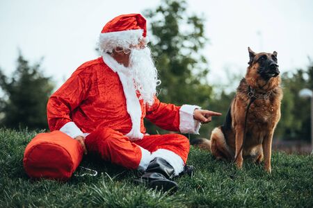 Stock photo of Santa claus sitting on the lawn, pointing to a dog with his finger and talking to him. Christmas time and Noelの写真素材