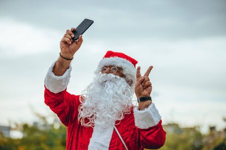 Stock photo of Santa Claus making a selfie with the mobile in one hand and the other making the symbol of victory. Christmas timeの写真素材