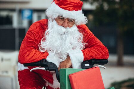Vertical stock photo of santa claus supported in a bicycle full of gifts with exhausted expression. Christmas timeの写真素材