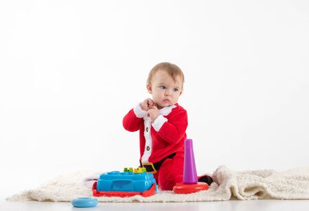 Stock studio photo with white background with a baby dressed as Santa Claus sitting on the floor playing with plastic toys.の写真素材