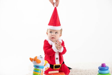 Stock studio photo of a baby disguised as Santa Claus sitting on the rolling floor of toys while one hand removes the hat from his head.の写真素材