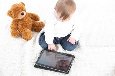Stock studio photo with a white background of a baby looking at the screen of a tablet with a teddy bear next to it.の写真素材
