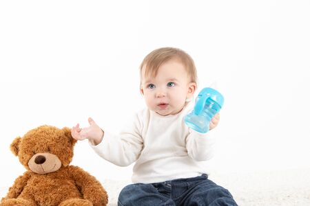 Stock studio photo with a white background of a baby with a canteen in his hand and a teddy bear by his side.の写真素材