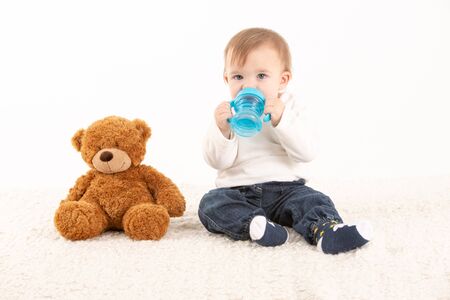 Stock studio photo with a white background of a baby with a canteen in his hand and a teddy bear by his side.の写真素材