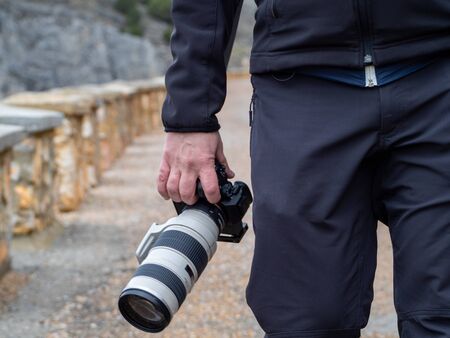 Hand of a man holding a camera with a big zoom objective outdoors in the middle of a path surrounded...