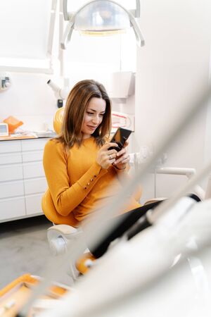Young female smiling and using smartphone while sitting on chair and waiting for doctor in contemporary dentist officeの写真素材
