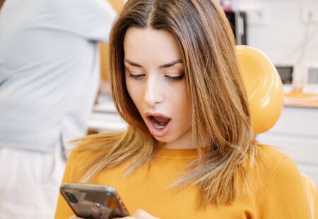 Astonished female patient sitting on chair and reading shocking news on smartphone while visiting dentist in clinicの写真素材
