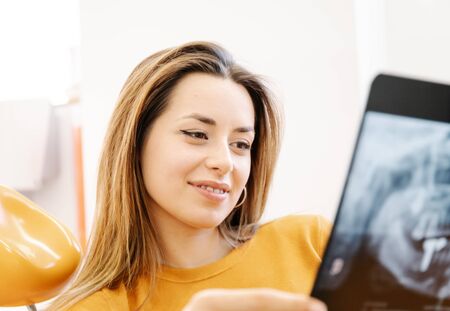 Young woman smiling and examining teeth X ray while sitting on chair in office of dental clinicの写真素材