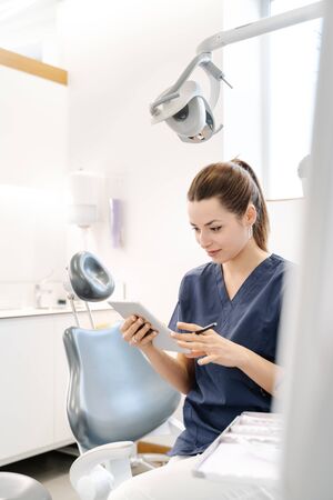 Young nurse wearing a blue jacket smiles in the dental office before receiving the patient and handles a tabletの写真素材