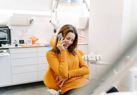 Young woman in orange sweater talks on the phone while waiting to be seen by the dentistの写真素材