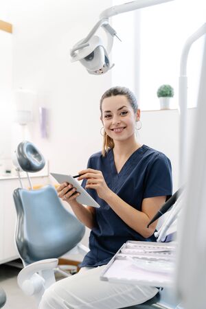 Young nurse wearing a blue jacket smiles in the dental office before receiving the patient and handles a tabletの写真素材