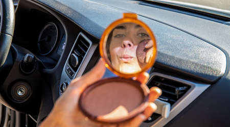 Crop female with pocket mirror pouting lips and applying rouge on cheek while sitting on passenger seat of vehicleの写真素材