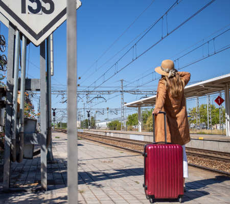 Back view of anonymous woman with suitcase waving hand with hat and walking on platform of railroad station during summer vacationの写真素材