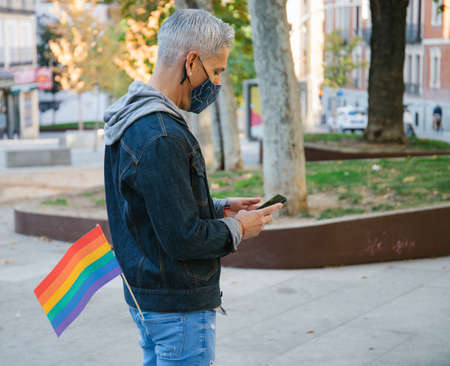 Side view of calm male in mask and with LGBT rainbow flag standing in street in Madrid and browsing cellphone during coronavirus pandemicの写真素材