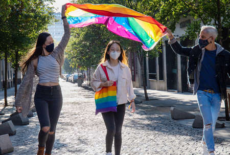 Group of cheerful multiethnic people in protective masks walking along street in Madrid with colorful LGBT flag and rainbow bagの写真素材