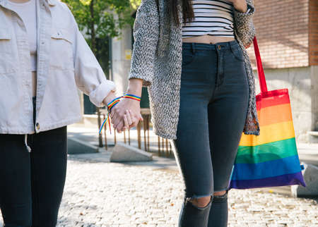 Crop faceless lesbian females with rainbow bag and bracelets holding hands and walking along street in cityの写真素材