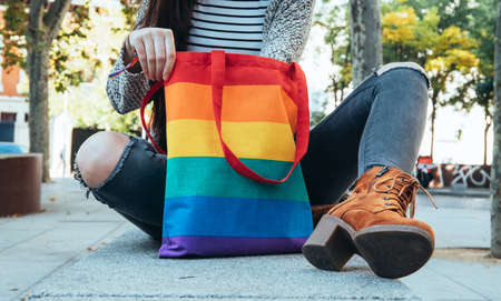 Calm female in protective mask sitting on pavement with rainbow LGBT shopping bag during coronavirus...