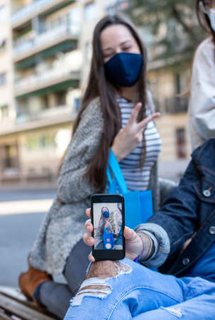 Group of diverse friends in protective masks and with paper bags taking selfie on smartphone after shopping in Madrid during coronavirus epidemicの写真素材