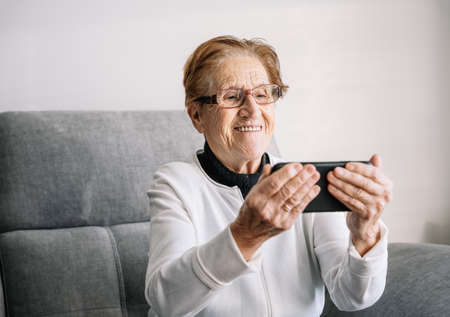 Cheerful aged female sitting on couch at home and speaking on video chat with family on mobile phone