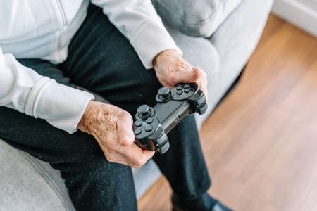 Crop anonymous 90 year old female sitting on couch with modern controller and playing video game at homeの写真素材