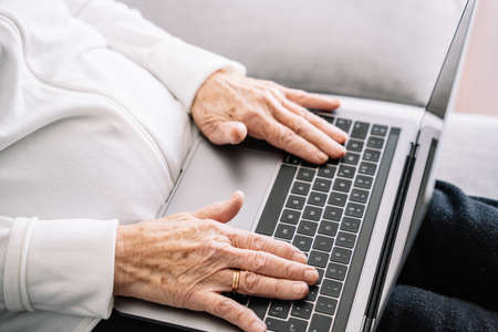 High angle of crop anonymous senior female sitting on sofa and browsing netbook while relaxing at home at weekendの写真素材