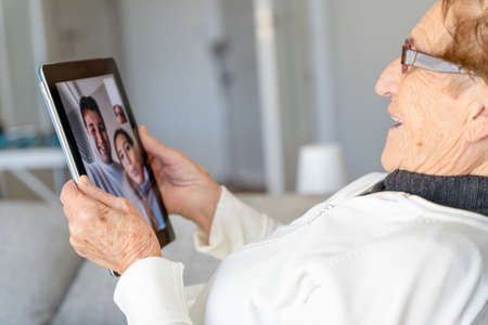 High angle side view of satisfied senior female resting on sofa at home and having video call with family while using tableの写真素材