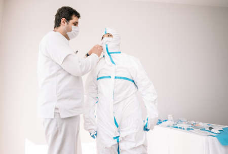 Medic in mask putting on protective costume with help of male coworker while standing in medical room during COVID 19の写真素材