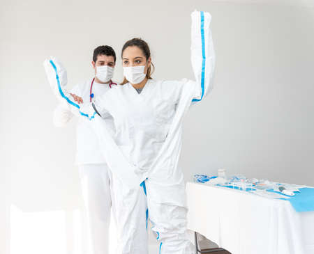 Medic helping female doctor in mask with putting on protective costume while standing in white medical room during coronavirus pandemicの写真素材