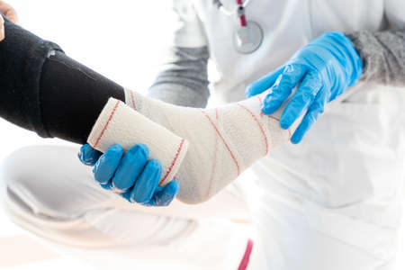 Crop medic in latex gloves wrapping leg of unrecognizable senior patient while using bandageの写真素材