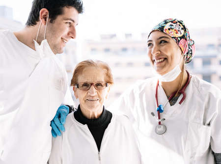 Delighted doctors hugging elderly female patient while sitting in room with sunlight and smilingの写真素材