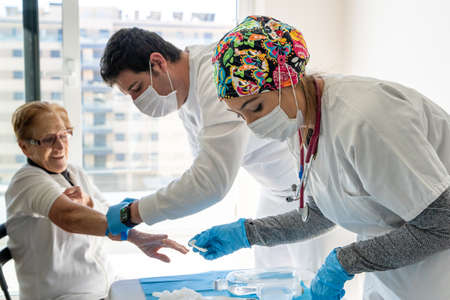 Side view of doctors in protective masks taking blood samples from elderly female sitting in room at homeの写真素材