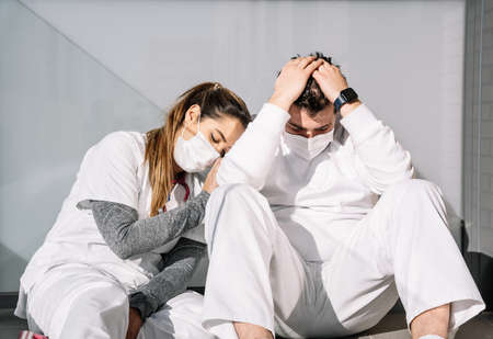 Exhausted male and female doctors in protective masks leaning on wall of terrace and sleeping after hard work during coronavirus epidemicの写真素材