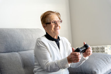 Glad senior female sitting on comfortable sofa in living room and playing video game while using gamepadの写真素材