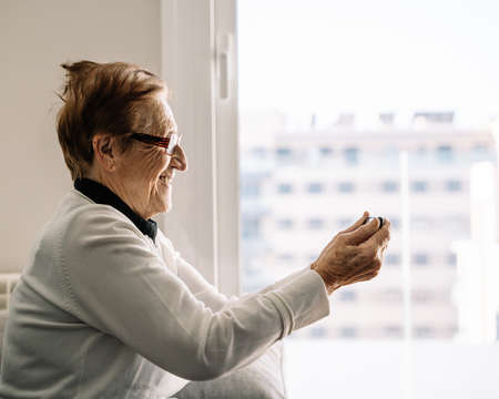 Side view of glad senior female having video chat with family on smartphone while sitting in living room at homeの写真素材