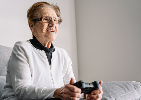 Crop anonymous 90 year old female sitting on couch with modern controller and playing video game at...