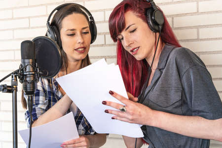 Young women in headphones talking about script while standing near microphone before radio program broadcast in studioの写真素材