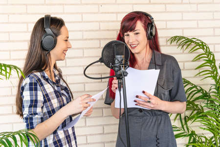 Happy young women in casual clothes and headphones smiling and talking with each other during radio program against brick wallの写真素材