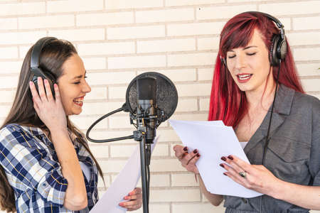Happy young voice actresses in headphones smiling and reading script near microphone while creating soundtrack for movie against brick wallの写真素材