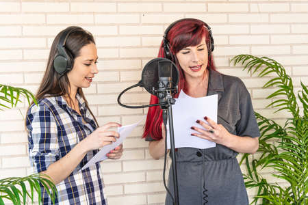Young women in headphones reading script from papers while standing near microphone and dubbing movie in studioの写真素材