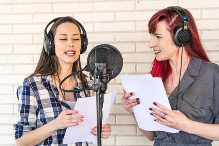 Happy young voice actresses in headphones smiling and reading script near microphone while creating soundtrack for movie against brick wallの写真素材