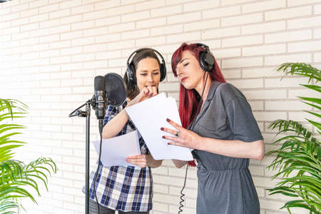 Young women in headphones discussing script while standing near microphone against brick wall in film dubbing studioの写真素材