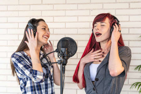 Happy young female in headphones smiling and listening to girlfriend singing song with closed eyes while recording music against brick wall in studioの写真素材