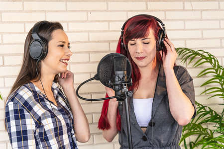 Happy young female in headphones smiling and listening to girlfriend singing song with closed eyes while recording music against brick wall in studioの写真素材