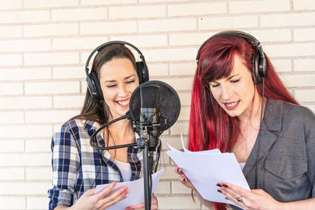 Happy young voice actresses in headphones smiling and reading script near microphone while creating soundtrack for movie against brick wallの写真素材