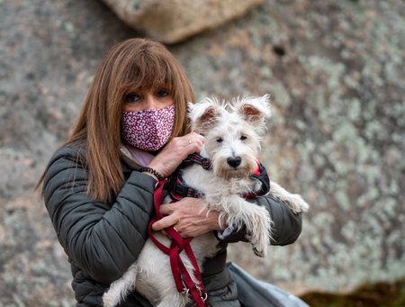 Adult woman with face mask holding white dog in a defocused stoned background outside.の写真素材