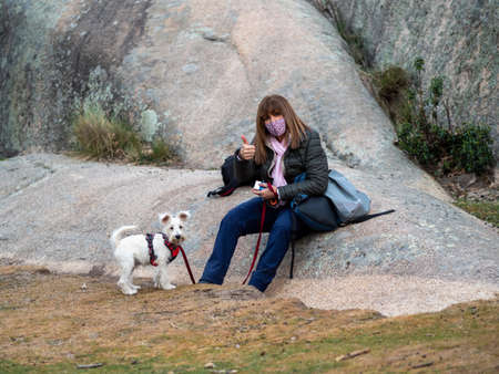 Adult woman sitting on a rock with her thumb up and her dog next to her, both looking at the camera. Woman wearing face mask and holding by leash her white dog.の写真素材