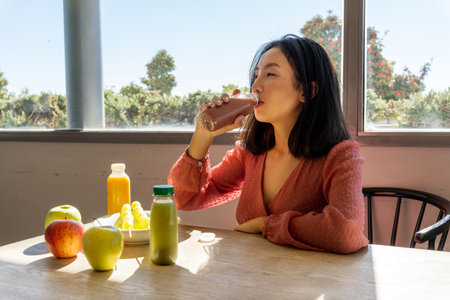 Asian woman drinking a healthy fruit and vegetable smoothie, sitting next to more fruits and juicesの写真素材