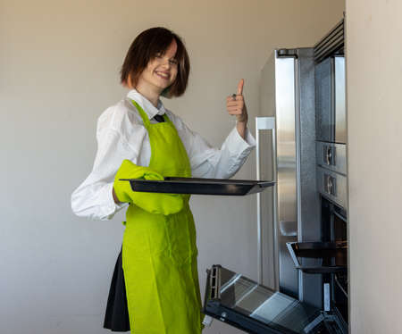 woman standing in green apron holding a baking tray in one hand and making an okay sign with the other. Plain backgroundの写真素材