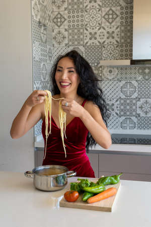 happy woman picking up cooked pasta with her hands, vegetables on a wooden board on the sideの写真素材