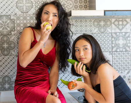 two well-dressed Asian girls posing in the kitchen with vegetables in their hands, tiled backgroundの写真素材